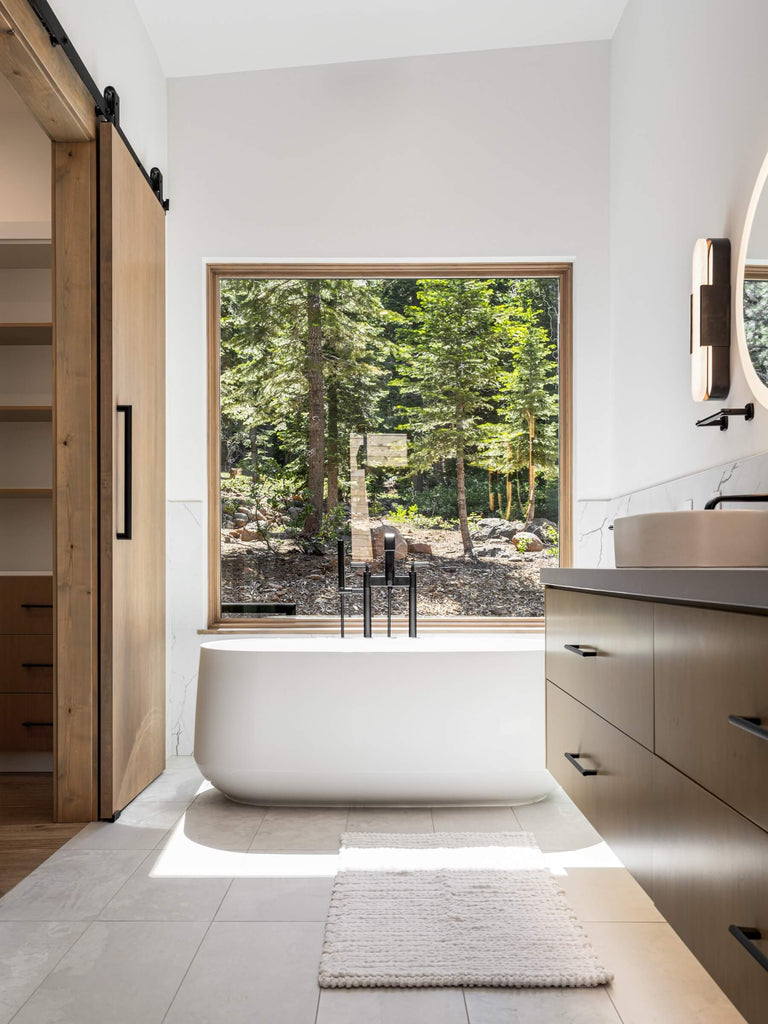 Contemporary bathroom with a freestanding bathtub, wooden cabinets, and a large window overlooking trees.